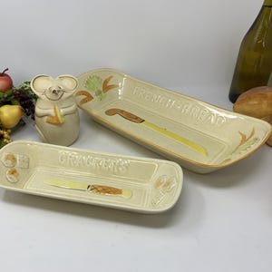Op de afbeelding: Twee rechthoekige keramische serveerschalen, een met het opschrift "FRENCH BREAD" en de andere "CRACKERS", met mesillustraties. Een keramisch muisbeeldje en een decoratief arrangement van fruit zijn ook aanwezig. Een groene glazen fles staat op de achtergrond.