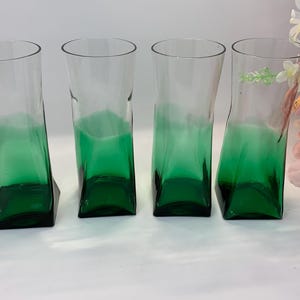 Op de afbeelding: Vier heldere glazen bekers met een groen ombre-effect. De glazen hebben een uniek, licht schuin ontwerp en een vierkante basis. De groene kleur is geconcentreerd aan de onderkant en vervaagt naar boven. De glazen zijn leeg.