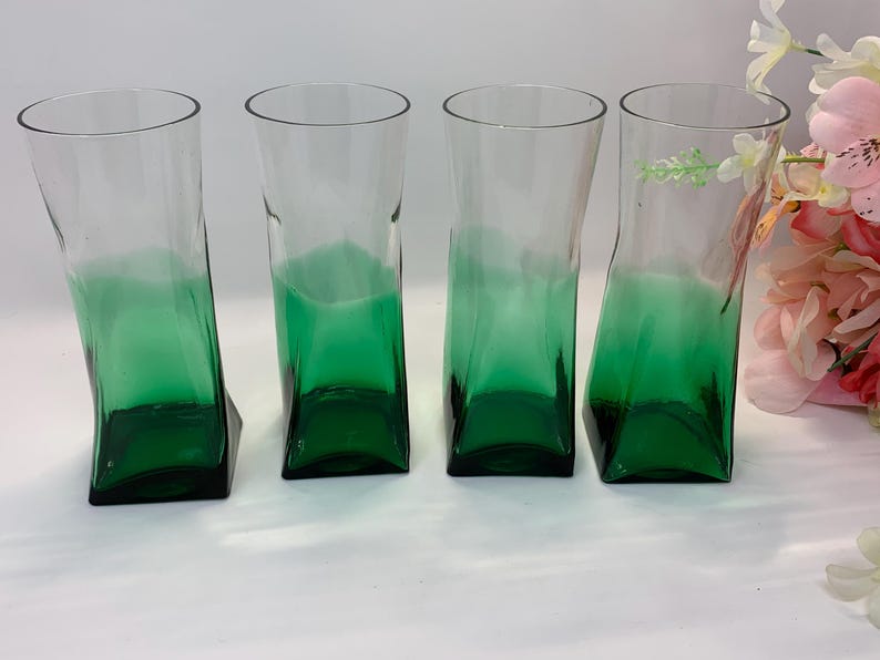 Op de afbeelding: Vier heldere glazen bekers met een groen ombre-effect. De glazen hebben een uniek, licht schuin ontwerp en een vierkante basis. De groene kleur is geconcentreerd aan de onderkant en vervaagt naar boven. De glazen zijn leeg.