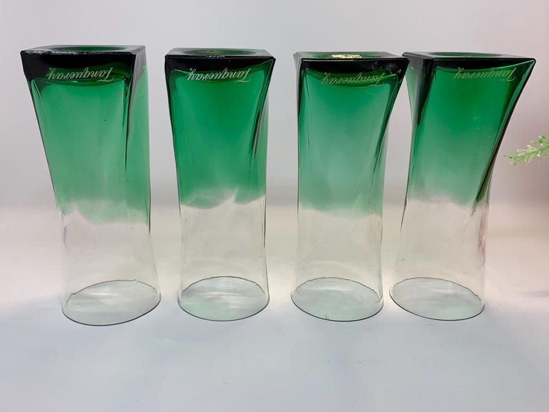 Op de afbeelding: Vier heldere, groen getinte glazen bekers met een vierkante bovenkant en gebogen zijkanten. De bovenste rand is zwart en het woord "Tanqueray" is in het wit gedrukt. De glazen hebben een gradi&euml;nteffect, van groen naar helder.