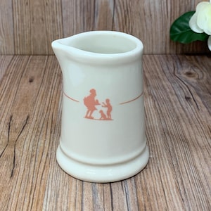 May include: A white ceramic creamer with a pink silhouette of two people and a dog. The creamer has a rounded body and a small spout.