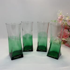 Op de afbeelding: Vier hoge, heldere drinkglazen met een groen ombre-effect aan de basis. De glazen hebben een geometrisch ontwerp en het woord "Tanqueray" is op de basis gedrukt. Een boeket roze en witte bloemen staat op de achtergrond.