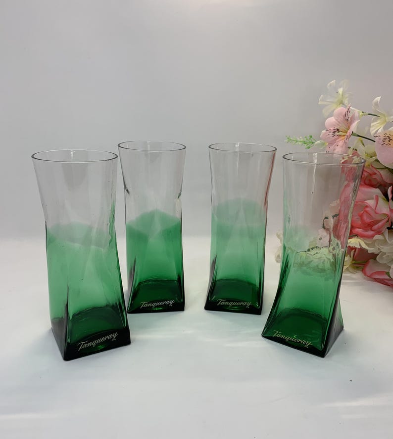 Op de afbeelding: Vier hoge, heldere drinkglazen met een groen ombre-effect aan de basis. De glazen hebben een geometrisch ontwerp en het woord "Tanqueray" is op de basis gedrukt. Een boeket roze en witte bloemen staat op de achtergrond.