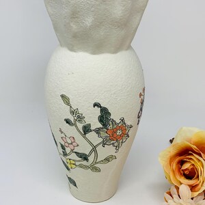 May include: Off-white ceramic vase with a textured surface and a flared, ruffled top. The vase is adorned with floral illustrations in shades of orange, pink, and green. A decorative piece for home decor.