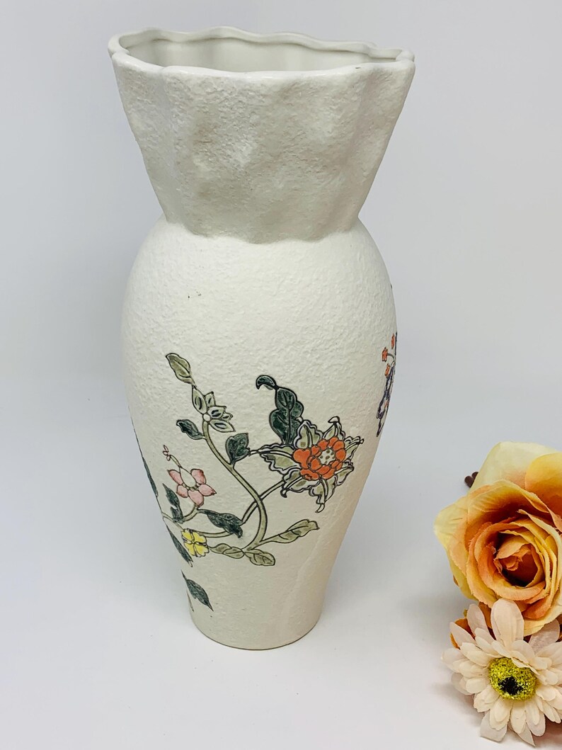 May include: Off-white ceramic vase with a textured surface and a flared, ruffled top. The vase is adorned with floral illustrations in shades of orange, pink, and green. A decorative piece for home decor.