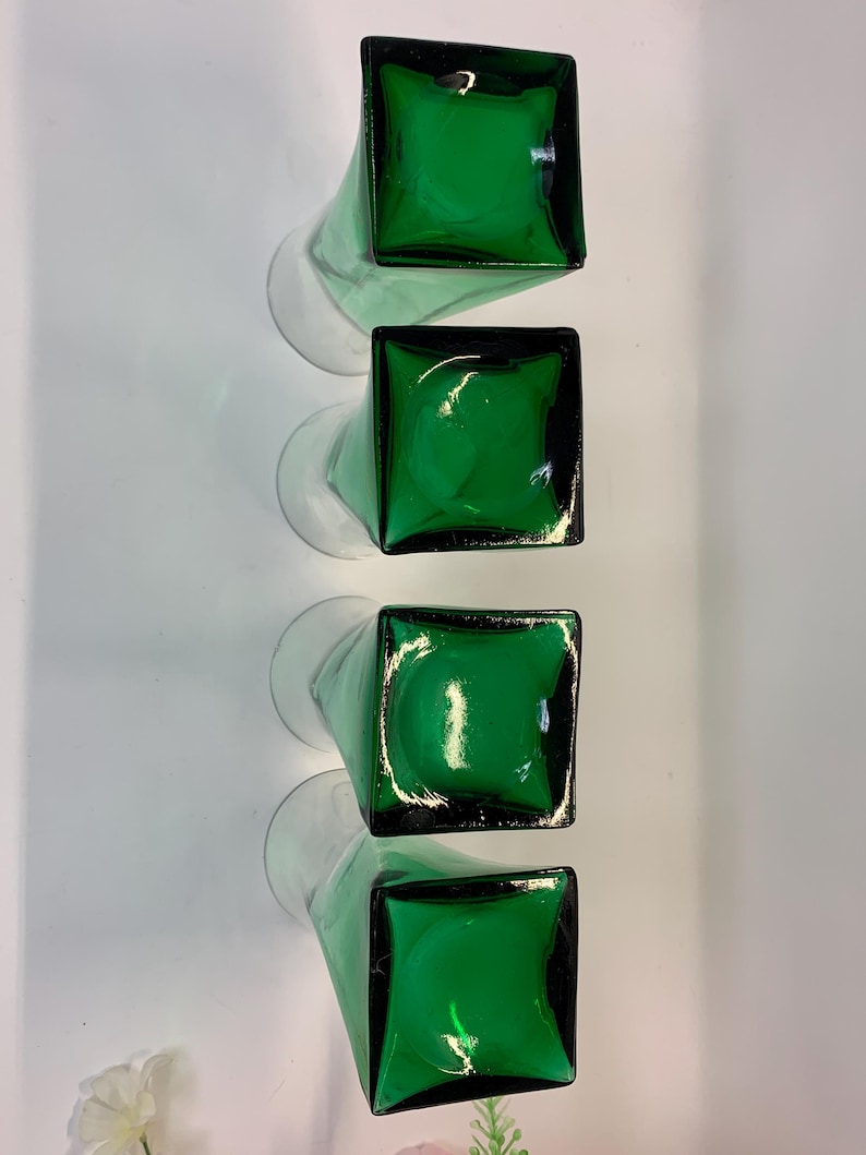 Op de afbeelding: Vier groene glazen vazen met vierkante bovenkanten en lange, heldere stelen zijn verticaal gerangschikt. De vazen hebben een glanzende afwerking en de groene kleur is levendig. De heldere stelen cre&euml;ren een contrast.
