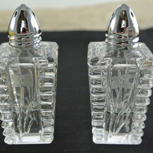 Puede incluir: Un juego de dos saleros y pimenteros de vidrio transparente con tapas plateadas. Los saleros tienen un diseño texturizado, geométrico y un grabado floral.