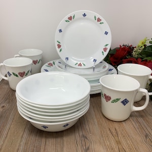 Puede incluir: Un juego de vajilla blanco con platos, cuencos y tazas. Los platos y tazas tienen un borde decorativo de corazones rojos, diamantes azules y hojas verdes. Los cuencos son blancos lisos.