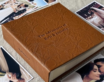 May include: A brown leather photo album with embossed text reading "The Adventures of Kris & Isabella" and "Volume 1." Several printed photographs of a wedding are scattered around the album.