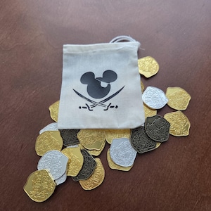 May include: A collection of pirate-themed coins in gold, silver, and bronze tones, scattered around a small canvas bag. The bag features a black graphic of a mouse head with crossed swords, suggesting a pirate theme.