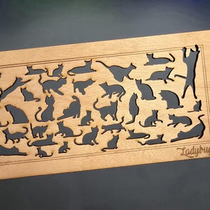 May include: A wooden plaque with a cutout design of multiple cats in various poses. The plaque is light brown and has the word "Ladybug" printed in the bottom right corner.