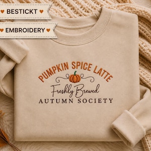 Könnte beinhalten: Ein beigefarbenes Sweatshirt mit Rundhalsausschnitt und einer Stickerei mit dem Text PUMPKIN SPICE LATTE, Freshly Brewed und AUTUMN SOCIETY, zentriert um eine kleine orangefarbene Kürbisgrafik mit grünem Stiel und schwarzen Schnörkeln.
