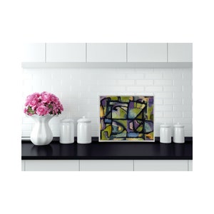May include: Abstract painting with geometric shapes in shades of green, purple, blue, and yellow. The painting is framed in a white frame and is hanging on a white tiled wall.