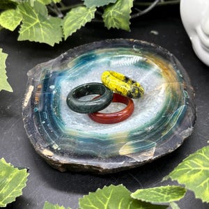 May include: A decorative agate stone bowl holds three rings: a dark green ring, a red ring, and a yellow ring with black spots. The agate bowl has blue, white, and brown bands. Green leaves surround the bowl.
