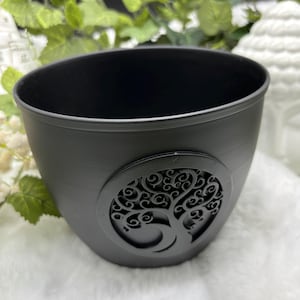 May include: A black, oval-shaped planter with a decorative tree of life design. The planter is made of a matte material and is suitable for indoor or outdoor use. The tree of life design is a dark gray color.