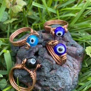 May include: Four copper wire wrapped rings with blue and black evil eye beads. The rings are displayed on a dark gray rock with green grass in the background.