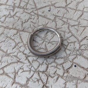 Op de afbeelding: Een zilverkleurige, scharnierende neusring met een spiraalvormig draadontwerp aan één kant. De ring is cirkelvormig en lijkt van metaal te zijn. De achtergrond is een getextureerd, gebroken wit oppervlak met een craquelé patroon.