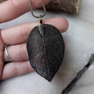 May include: A bronze-toned leaf pendant necklace. The large leaf-shaped pendant features intricate veining details. The necklace has a delicate chain and a small leaf accent at the top of the pendant. The overall design is nature-inspired.