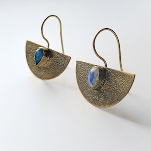 Può includere: Un paio di orecchini in ottone con un design a mezzaluna. Ogni orecchino presenta una superficie strutturata e una pietra preziosa blu a forma di goccia. Gli orecchini hanno una chiusura a gancio. Sono un gioiello.