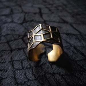 Puede incluir: Un anillo de oro con un diseño geométrico. El anillo está hecho de metal y tiene un acabado negro.