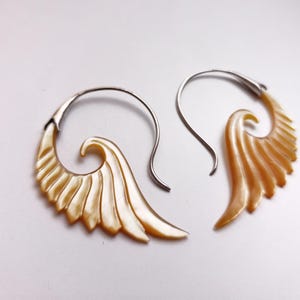Könnte beinhalten: Ein Paar Ohrringe mit einem flügelartigen Design. Die Ohrringe haben einen silbernen Haken und ein wirbelndes, fächerförmiges Element in einer hellen, perlmuttartigen Farbe. Das Design hat ein geschichtetes, strukturiertes Aussehen, das an Federn erinnert.