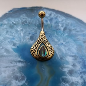 May include: A belly button ring featuring a teardrop pendant. The pendant has an ornate, gold-coloured filigree design with an iridescent centre. The top ball is gold-coloured, and the post is silver-coloured.