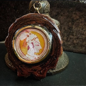 Op de afbeelding: Een ronde houten hanger met een gouden, ronde inzet met een geschilderd portret van een persoon met een hoofdtooi. De hanger heeft een bruin en gouden kleurenschema.