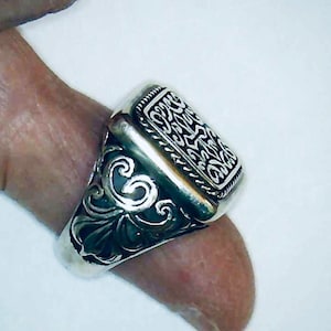 May include: A silver ring with a square-shaped top featuring an intricate, engraved design. The ring has a detailed, ornate band.