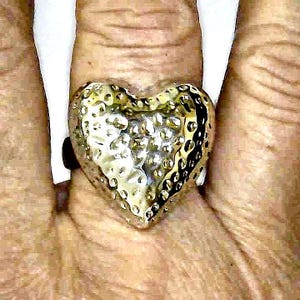 May include: A textured, gold-colored heart-shaped ring. The ring has a hammered appearance, with a slightly irregular surface. The heart shape is prominent, and the ring appears to be made of metal.