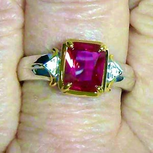 May include: A ring featuring a large, rectangular, vibrant pink gemstone set in a gold-toned prong setting. The band is silver-toned and includes two smaller, triangular-shaped clear stones on either side of the central pink gemstone.