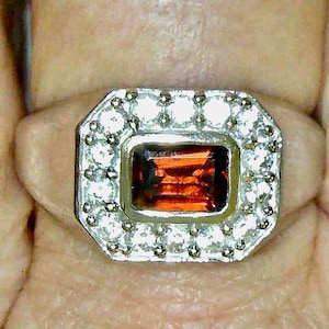 May include: A silver ring with a rectangular, orange-red gemstone at the center, surrounded by a halo of small, clear stones. The ring's setting has an octagonal shape.