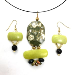 May include: A handmade jewelry set featuring a pendant and matching earrings. The pendant has a large, faceted green stone with a patterned design, complemented by a light green rectangular stone and smaller beads. The earrings mirror the design with light green stones and black beads.