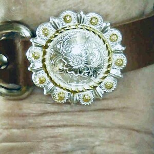 May include: A brown leather bracelet with a silver and gold concho. The concho has a floral design.