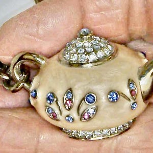 Puede incluir: Un charm de tetera blanca con una cadena dorada, decorado con diamantes de imitación azules y rosas. La tetera tiene una tapa con una parte superior de cristal y un asa detallada.