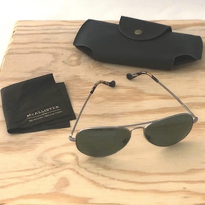 May include: Aviator sunglasses with dark green lenses and thin metal frames, featuring tortoise shell pattern temple tips. Includes a black protective case with a snap closure and a black microfiber cleaning cloth with the text McALLISTER.