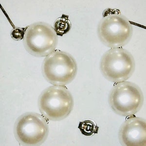May include: A pair of elegant, white pearl earrings. Each earring features a series of large, round pearls strung together, with a silver-toned post and backing. The pearls have a lustrous sheen, reflecting light. The earrings are displayed against a plain background.