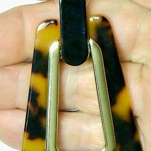 May include: A geometric earring with a tortoiseshell pattern, featuring a black top and a gold-colored metal frame. The earring has a rectangular top and a trapezoid shape.