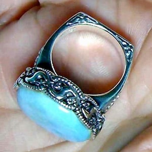 May include: A silver ring with a light blue oval stone. The ring has an ornate, detailed design around the stone and on the band. The ring is held in a hand.