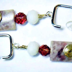 May include: A pair of handmade earrings featuring a square, light-colored stone with a gold spiral accent. The earrings also include a red faceted bead, a white bead, and a silver square post with a clear earring back.