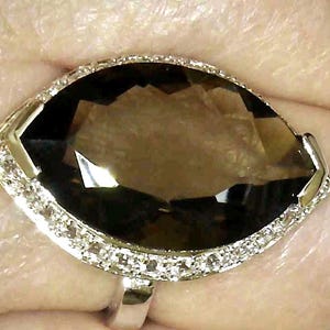 May include: A silver ring featuring a large, marquise-cut, smoky brown gemstone. The gemstone is surrounded by a row of small, clear accent stones. The ring is set against a skin-toned background.