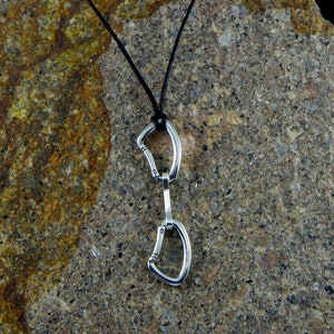 May include: A silver necklace featuring a double carabiner pendant. The pendant is made from two connected carabiners, a common climbing accessory. The necklace is on a black cord and is displayed against a rough, brown rock surface.