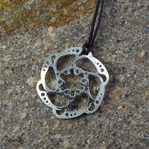 Peut inclure: Un collier avec un pendentif en argent de forme circulaire orné d'un motif détaillé et complexe. Le pendentif est attaché à un cordon noir.