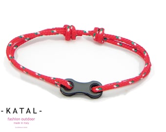 May include: Red and white rope bracelet with a black clasp. The bracelet is made of a red rope with white and green accents. The clasp is a black, stylized shape. The bracelet is a fashion accessory.