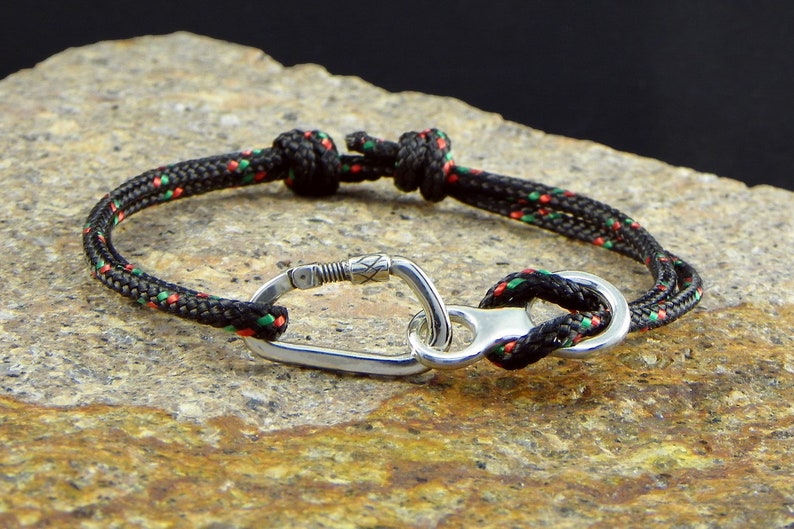 May include: A black and green rope bracelet with a silver carabiner clasp. The bracelet is made of a durable, braided rope and features a secure clasp.