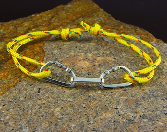 May include: A yellow rope bracelet with a silver carabiner clasp. The rope is accented with red and blue stripes.