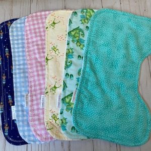 May include: A set of six contoured burp cloths featuring various patterns including navy blue with rockets, light blue gingham, pink gingham, yellow with green frogs, light blue with green dinosaurs, and a solid teal fabric with white dots.
