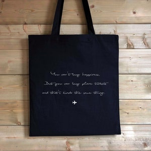 May include: Black tote bag with white text that reads "You can't buy happiness, But you can buy plane tickets and that's kinda the same thing."  A small airplane symbol is below the text.