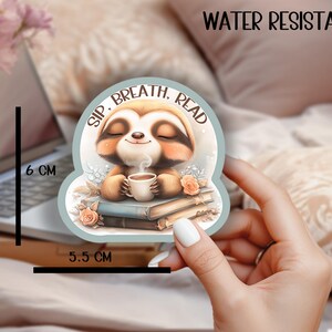 Könnte beinhalten: Wasserfester Aufkleber mit einem Cartoon-Faultier, das eine Tasse Kaffee hält und von Büchern umgeben ist. Der Aufkleber trägt die Aufschrift "Sip, Breath, Read" und misst 5,5 cm x 6 cm.