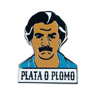 May include: A silver enamel pin featuring a cartoon illustration of a man with a moustache and the text "Plata o Plomo" in white letters on a black background.