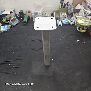 May include: A silver metal post with a square top and four holes. The post is rectangular and stands upright. The base is also rectangular. The post is likely for construction or furniture support.